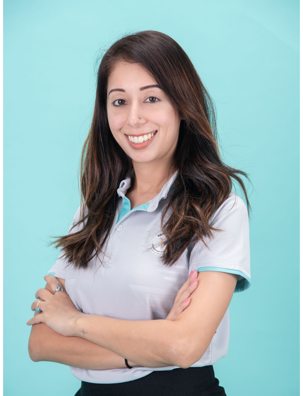Woman with crossed arms smiles, wearing a light blue and white shirt against a teal background.