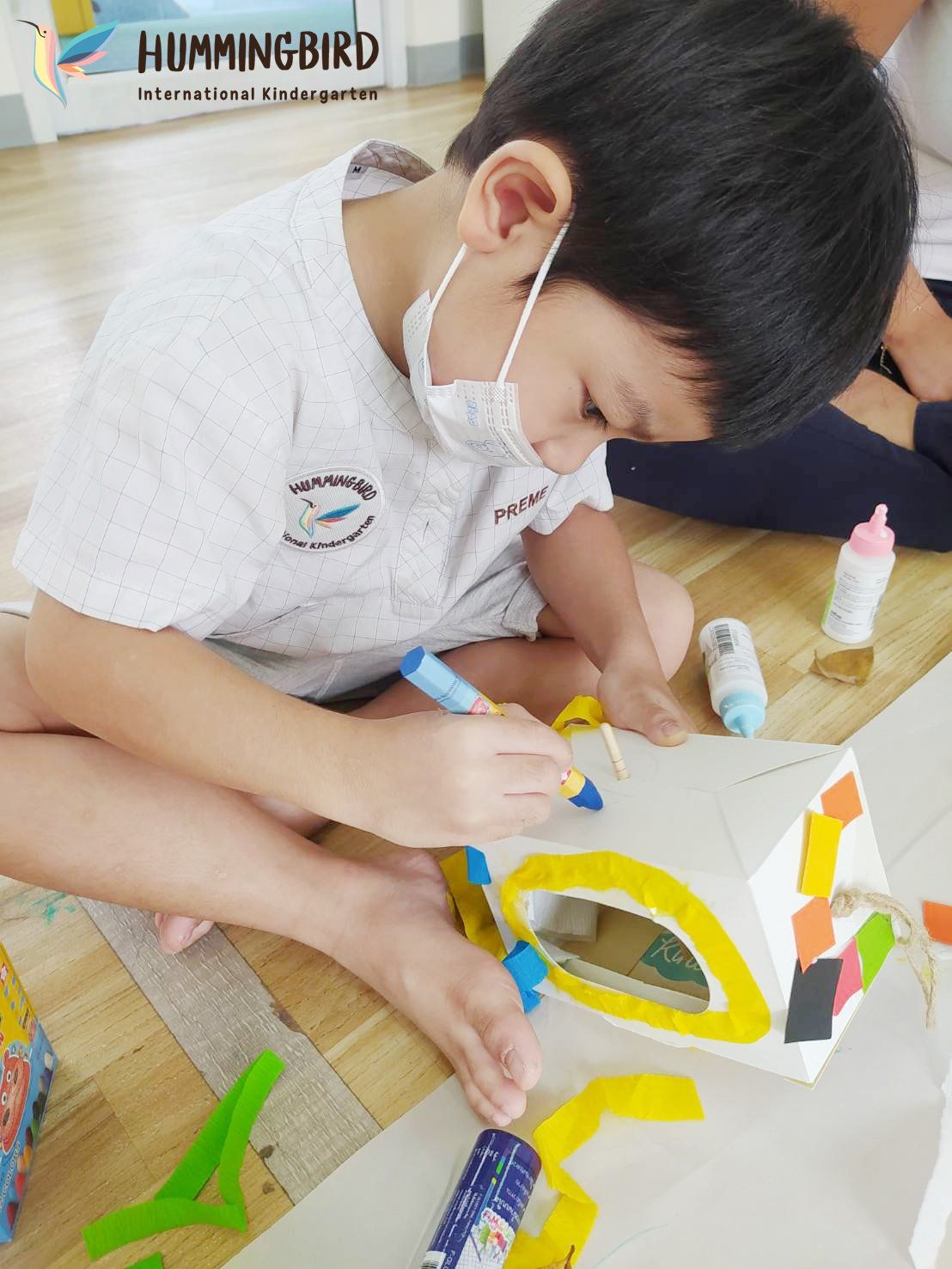 A young boy wearing a mask is sitting on the floor drawing on a box.