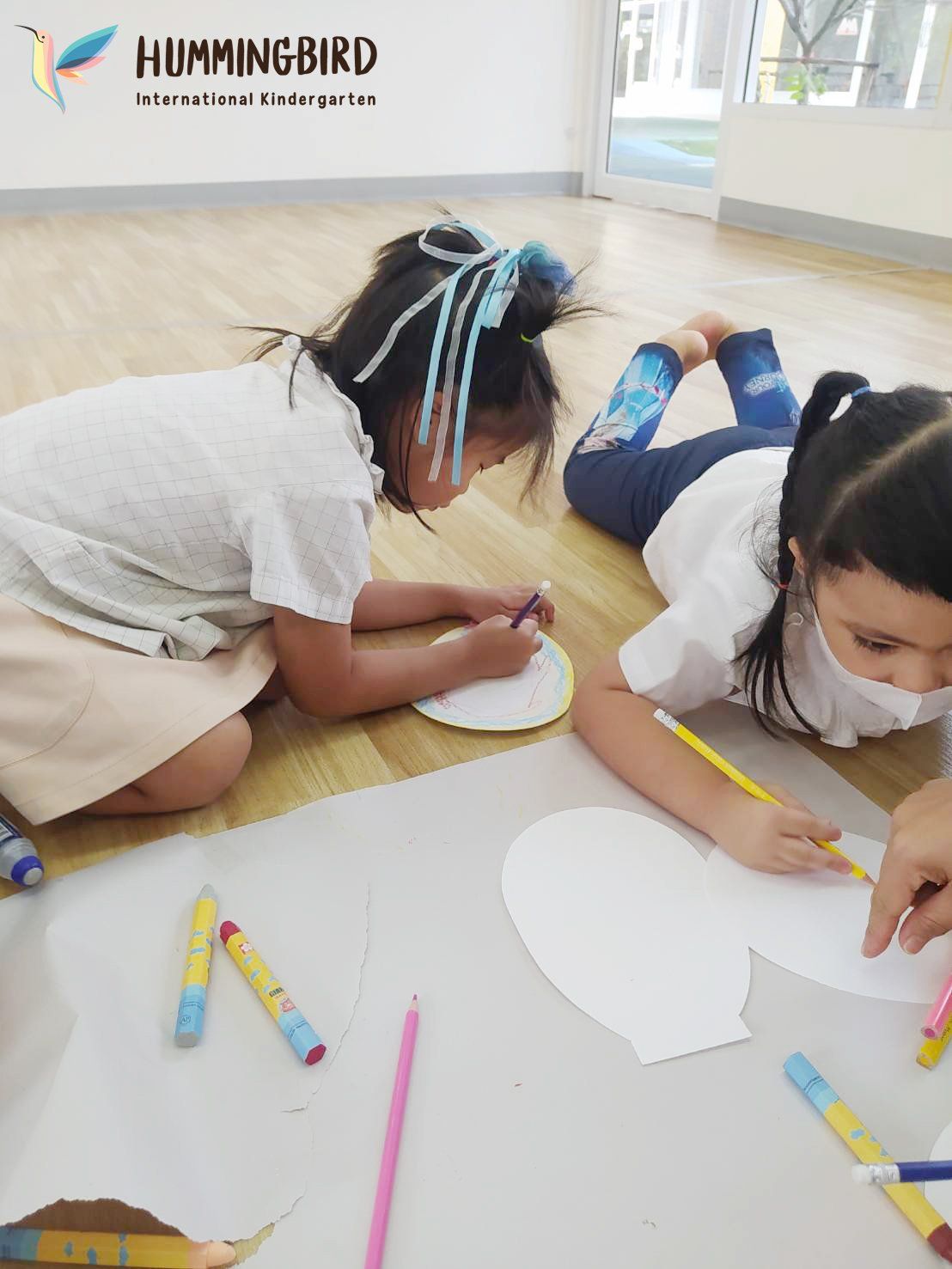 Two young girls are laying on the floor drawing on a piece of paper.