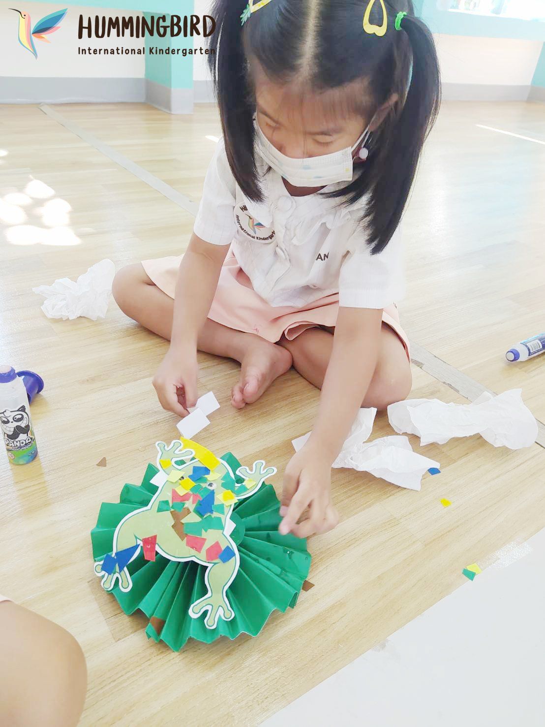 A little girl is sitting on the floor making a paper frog.