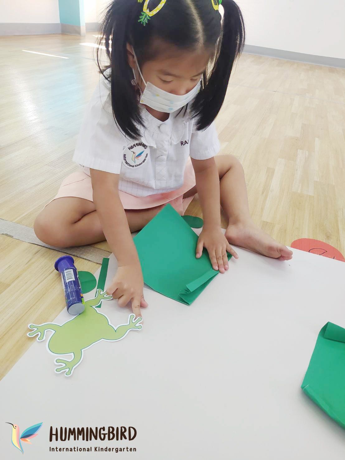 A little girl wearing a mask is sitting on the floor making a frog out of paper.