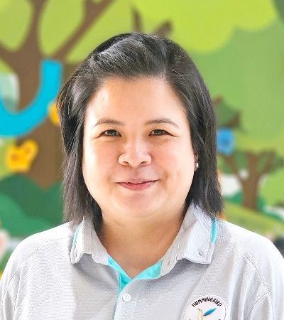 A woman in a grey shirt is smiling in front of a painting of trees.