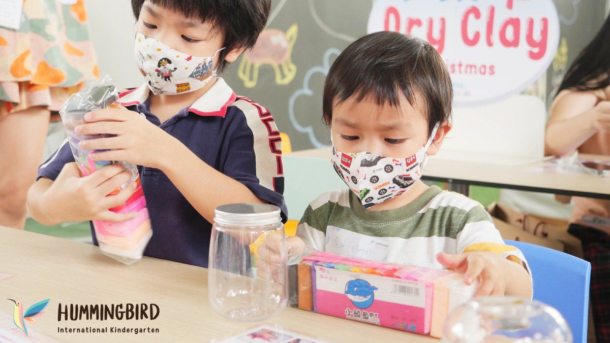 Two young boys wearing face masks are sitting at a table playing with dry clay.