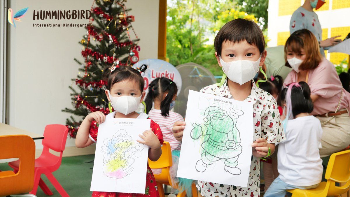 Two children wearing face masks are holding up their drawings in front of a christmas tree.