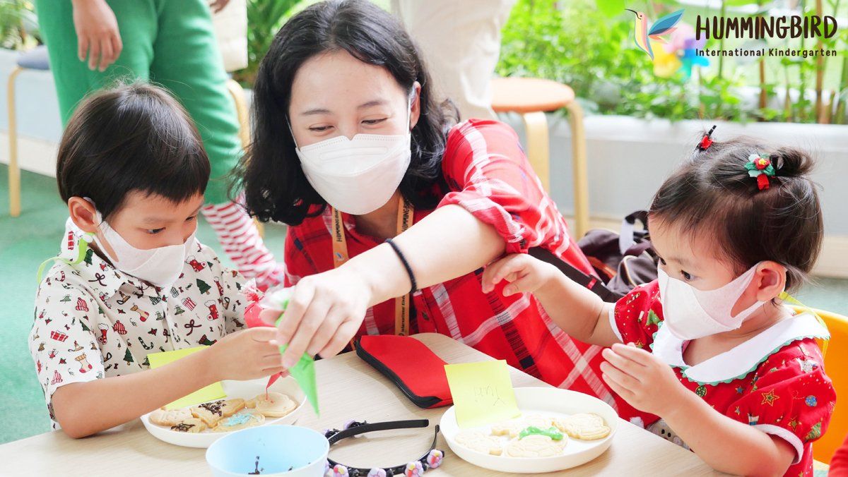 A woman wearing a mask is sitting at a table with two children.