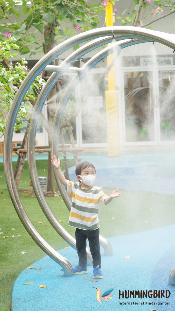A young boy wearing a mask is standing in front of a water fountain.