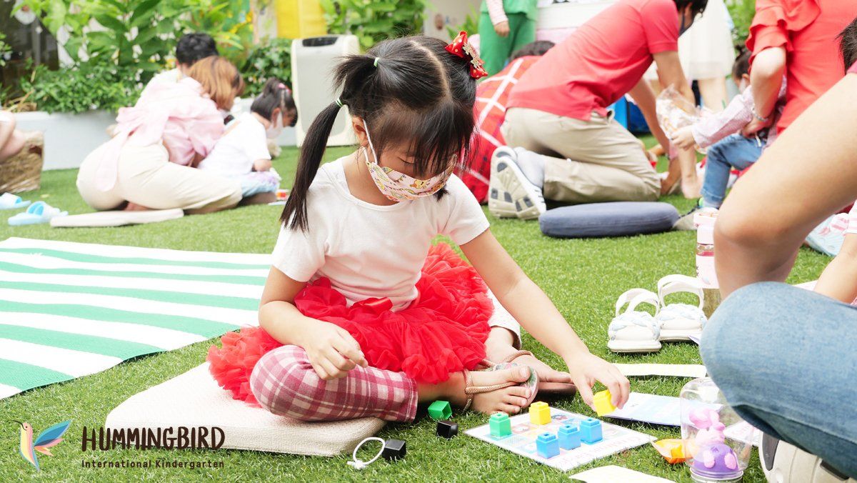 A little girl wearing a mask is sitting on the grass playing with toys.