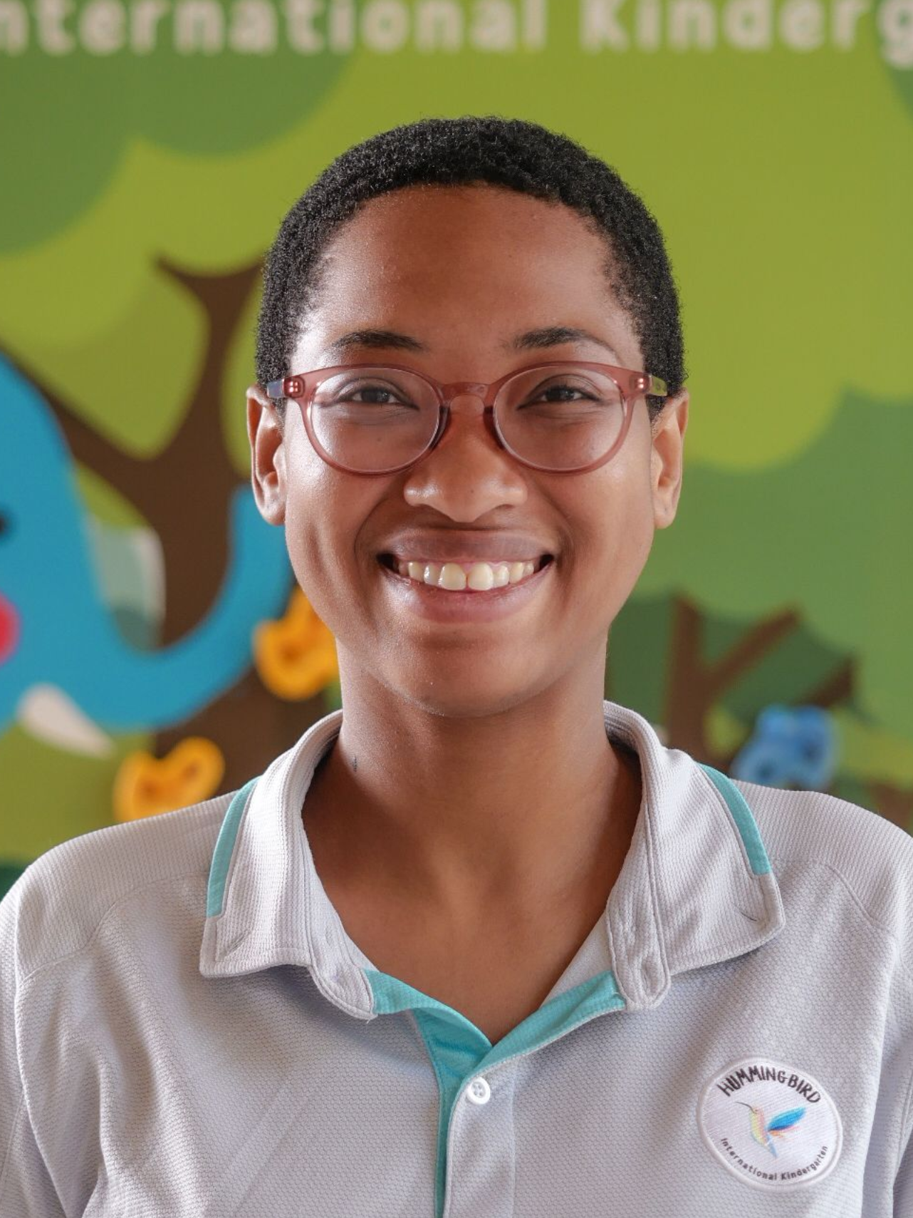 Woman with glasses smiling at the camera, wearing a light-blue collared shirt. Kindergarten backdrop.