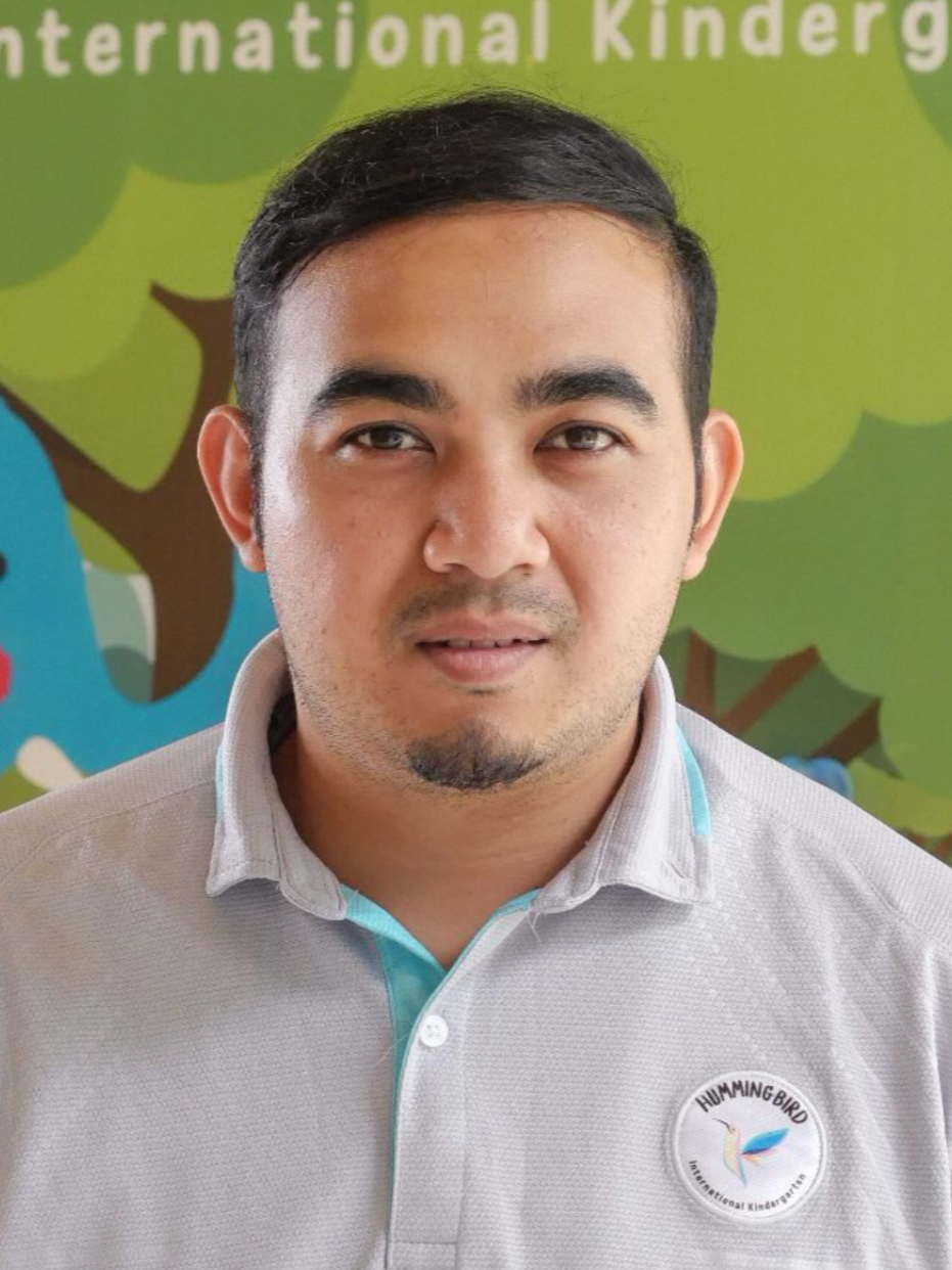 Man in gray polo shirt, smiling, in front of a kindergarten backdrop with a logo.