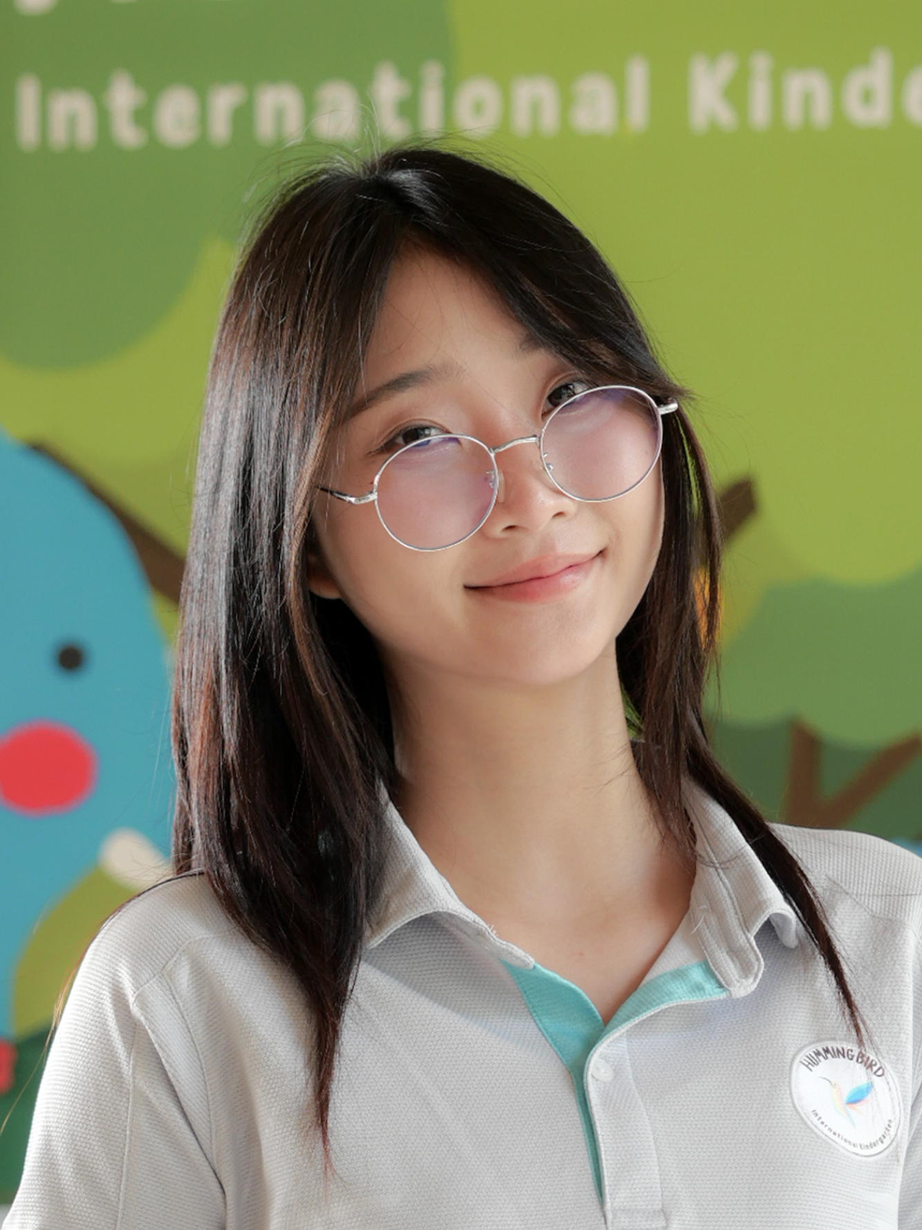 Woman with glasses, smiling, wearing a light gray polo shirt. Green and blue background.