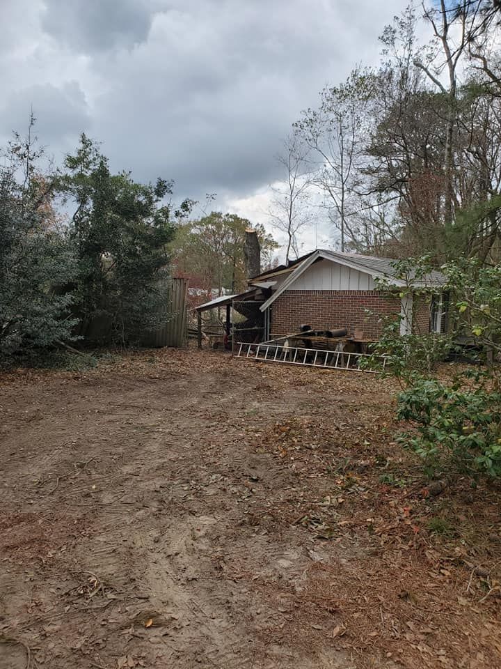 Exterior view of a dilapidated brick building with a missing roof, surrounded by trees and a dirt yard on an overcast day.
