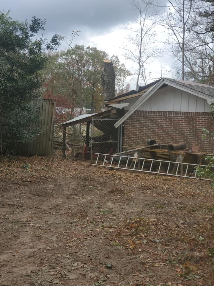 A brick building with a damaged roof and a leaning shed. A ladder sits in front of the building.
