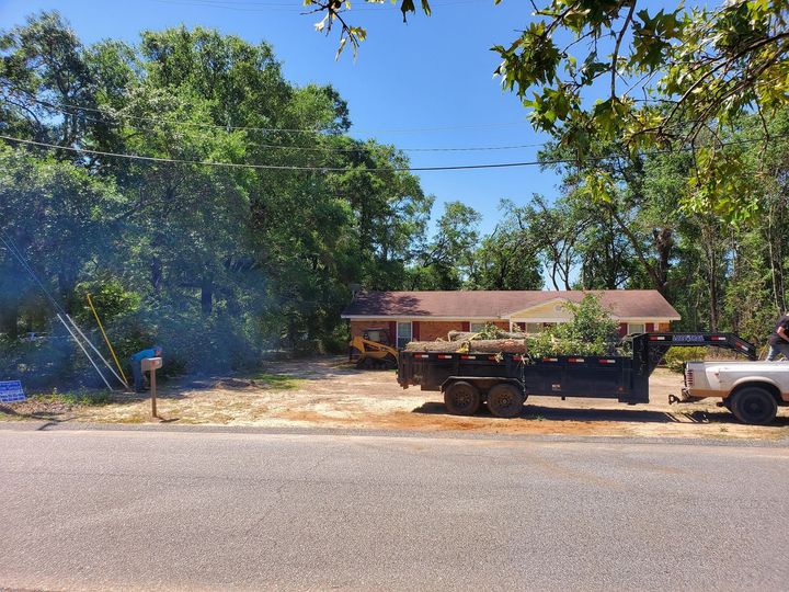 House with a trailer loaded with branches on the front lawn being worked on under a blue sky.