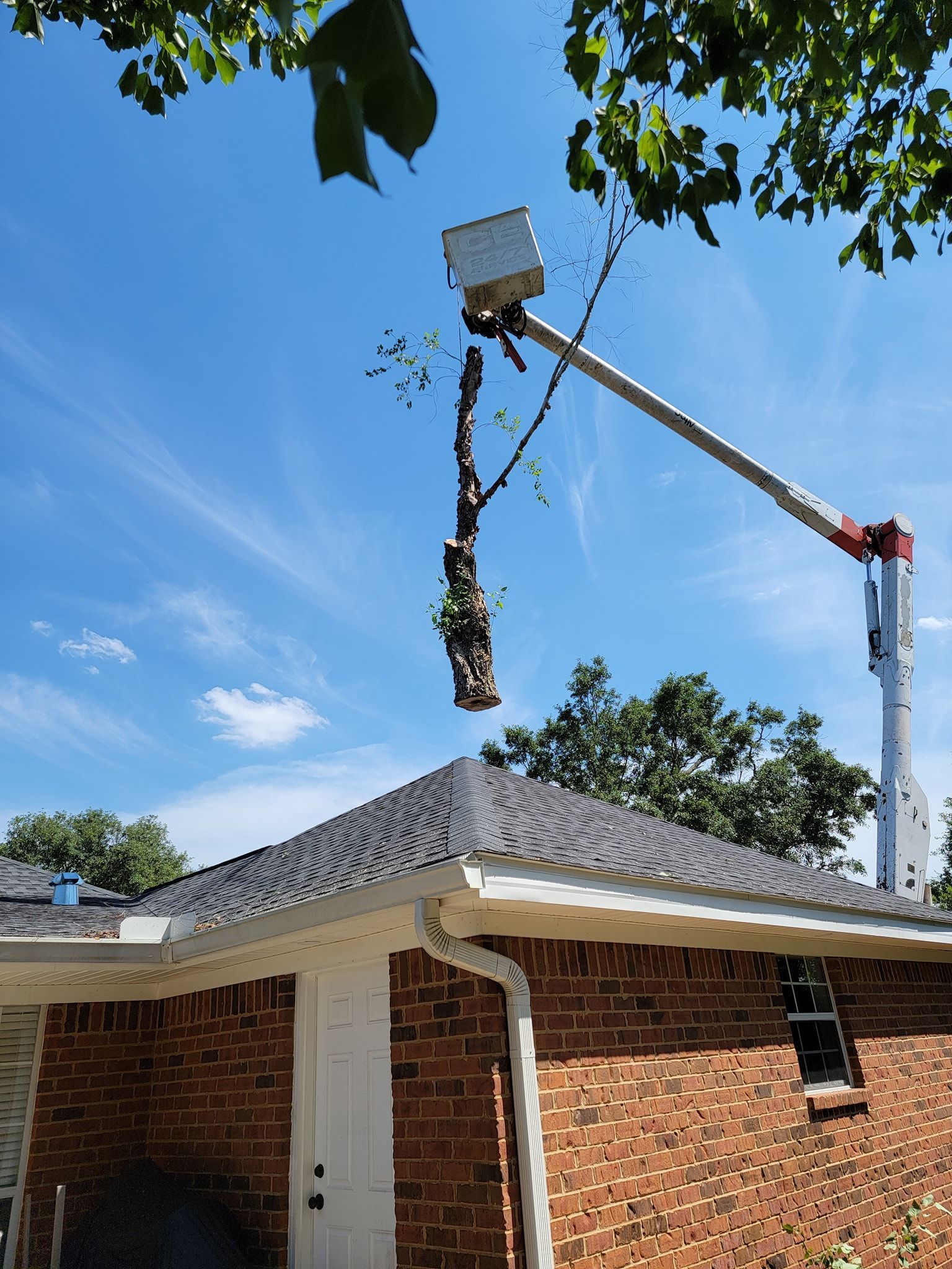 A tree being trimmed by a lift crane over a brick house with a dark roof on a sunny day.