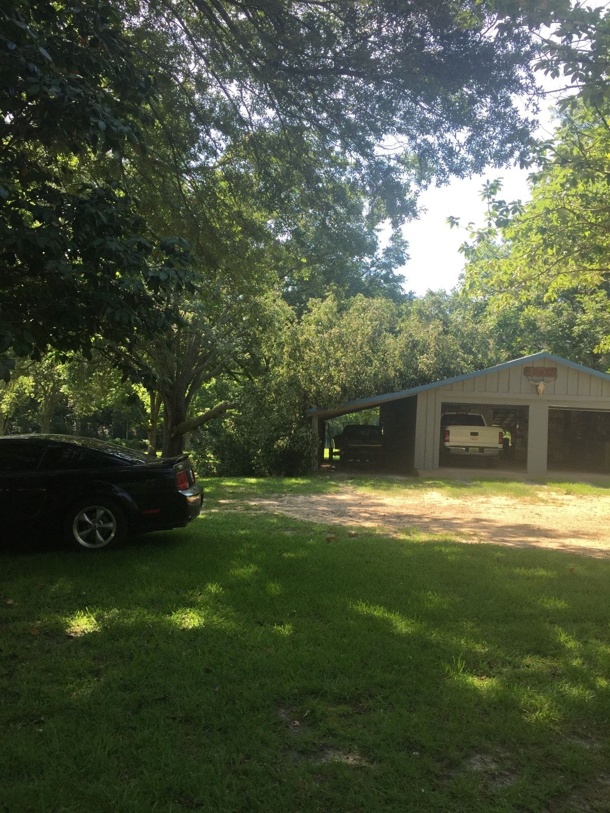 Black car parked on a grassy yard next to a two-bay garage under trees on a sunny day.
