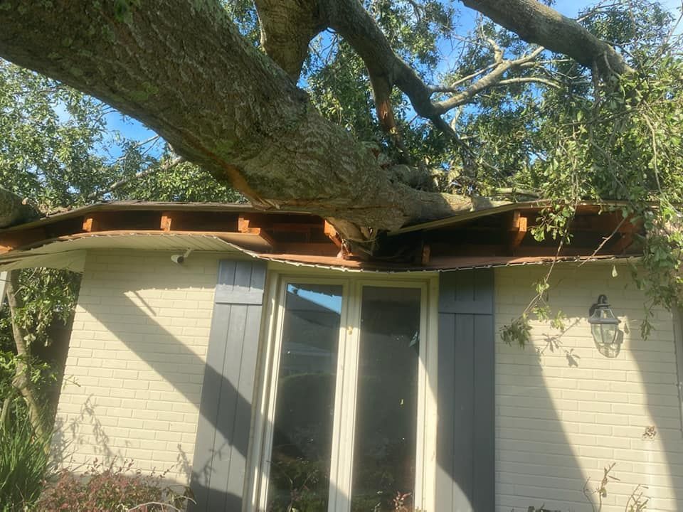 Tree fallen on a white brick house, crushing the roof above a window and door.