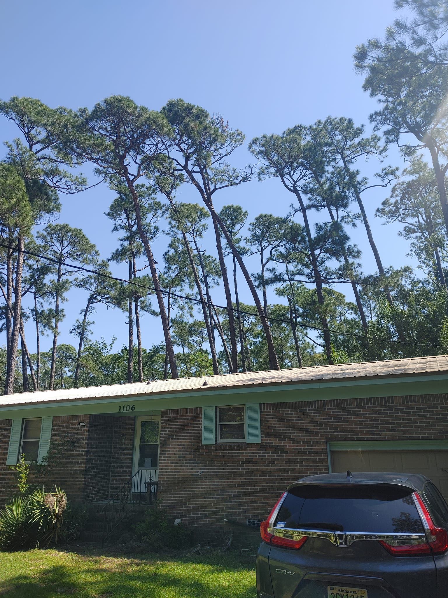 A single-story brick house with a blue car parked in front, tall trees in the background.