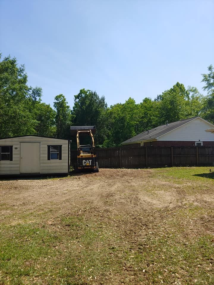 A yellow skid steer tractor is in a yard between a shed and a fence, clearing dirt under a blue sky.