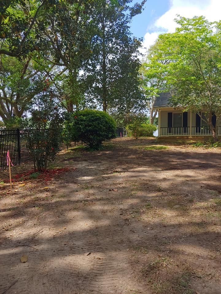 Dirt path leading to a white house with a porch, surrounded by trees and a black fence.