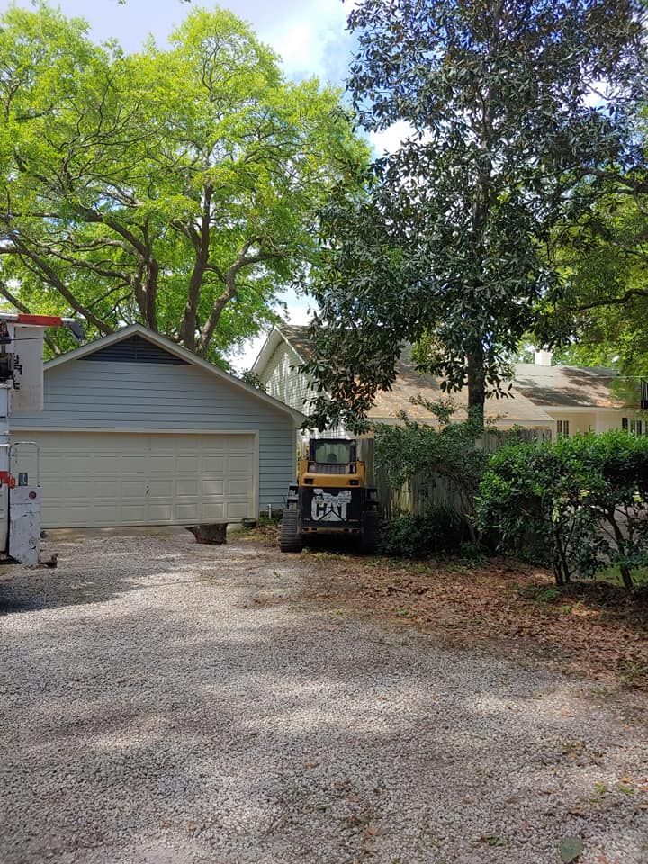 Blue garage with white door, gravel driveway, small yellow loader, lush green trees.
