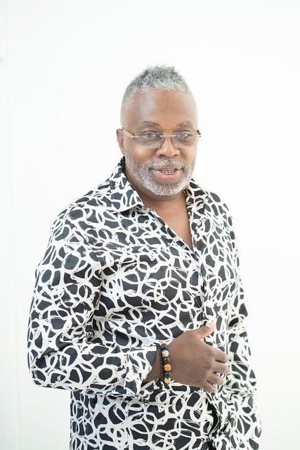 A person with short, graying hair and glasses wears a black and white patterned shirt, smiling against a white background.