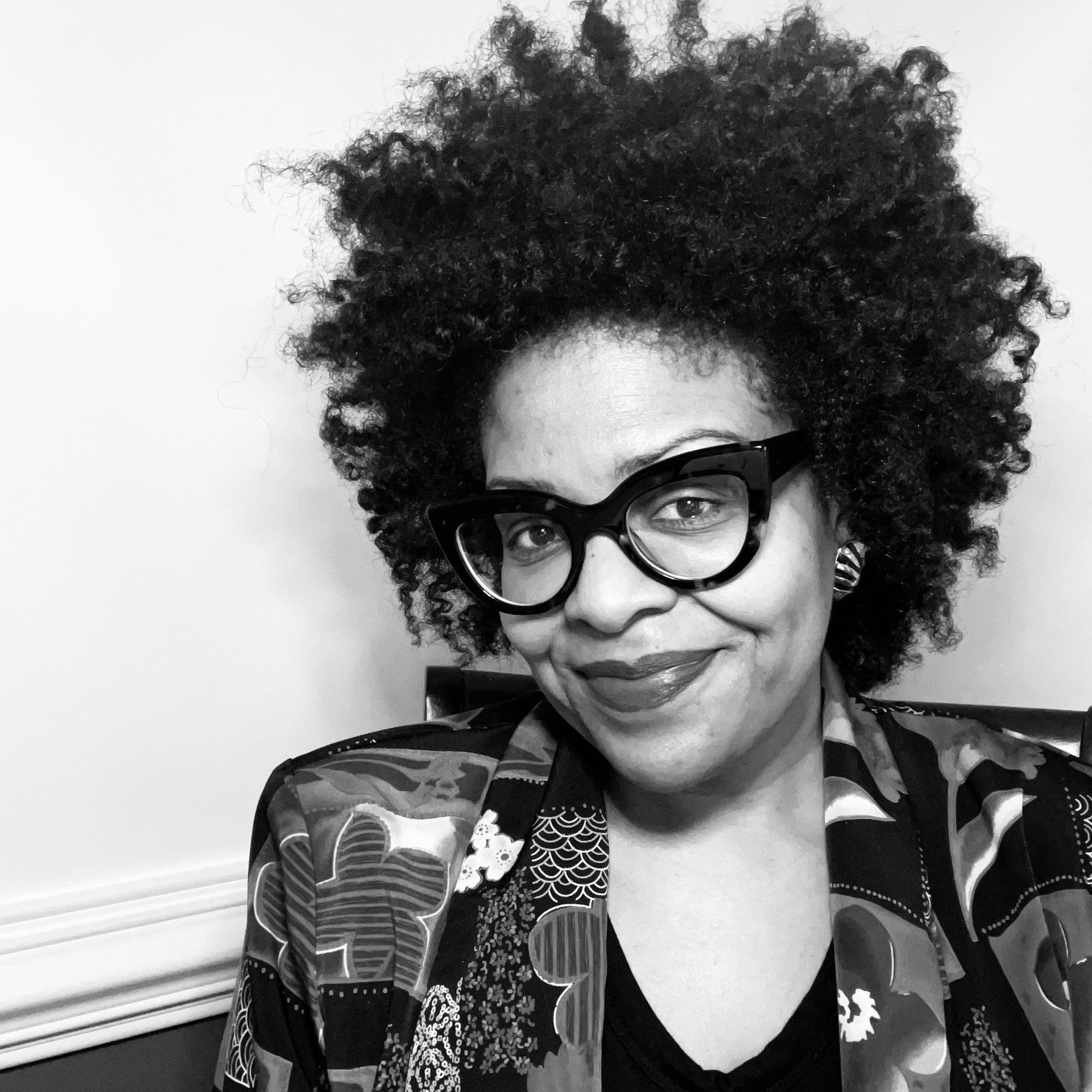 A person with curly hair and thick-rimmed glasses wearing a patterned blazer, looking directly at the camera.