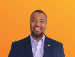 A person with a beard, wearing a blue blazer and light shirt, smiling in front of a bright orange background.