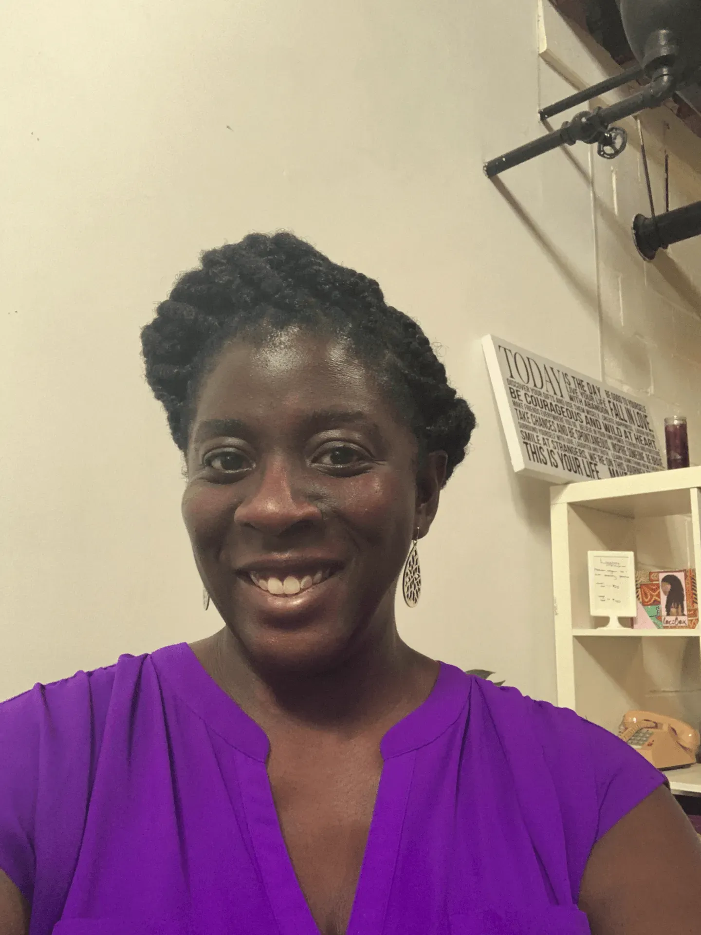 A person smiling, wearing a bright purple top with a pleated neckline, standing indoors in front of a white shelving unit.
