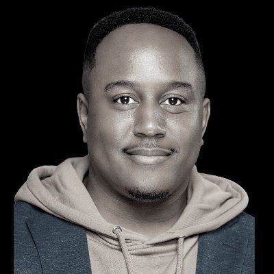 A studio portrait of a person smiling softly, wearing a hooded sweatshirt under a blazer against a plain black background.