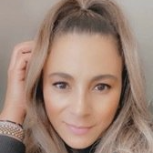 A person with shoulder-length, light brown hair styled in a half-up ponytail looks toward the camera with a subtle smile.