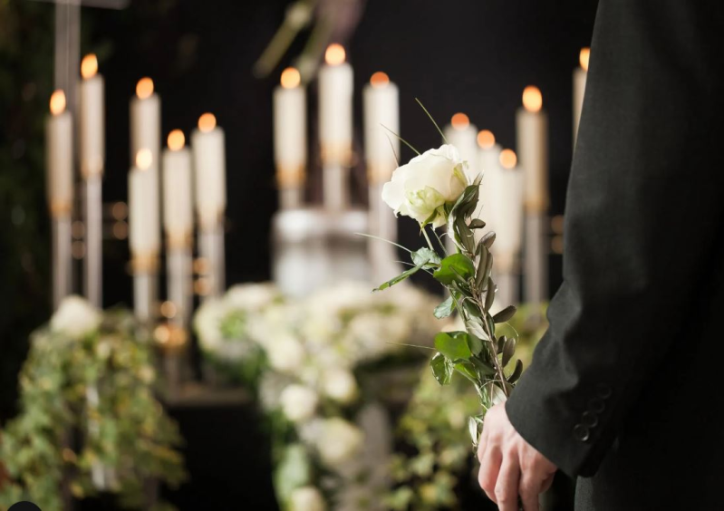 Persona che tiene in mano una rosa bianca durante un funerale, circondata da fiori e candele.
