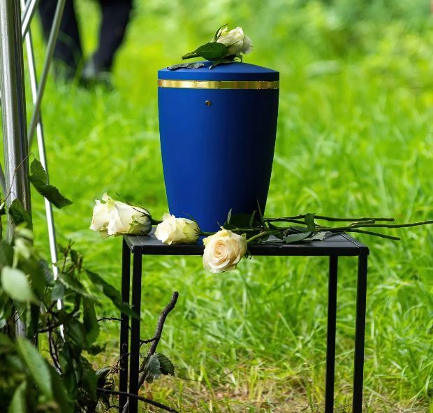 Urna blu con finiture dorate e rose bianche su un supporto nero, all'aperto sull'erba.