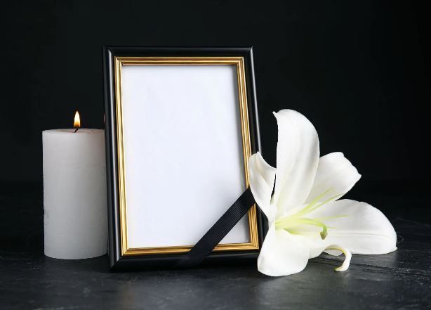 Foto con cornice nera, giglio bianco, candela accesa e nastro nero su superficie nera; commemorativo.