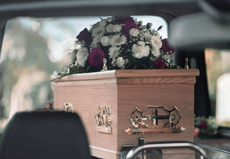 Una bara di legno con una composizione floreale all'interno di un veicolo, probabilmente un carro funebre.