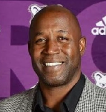 Smiling man in a gray checkered jacket at a purple event backdrop with logos