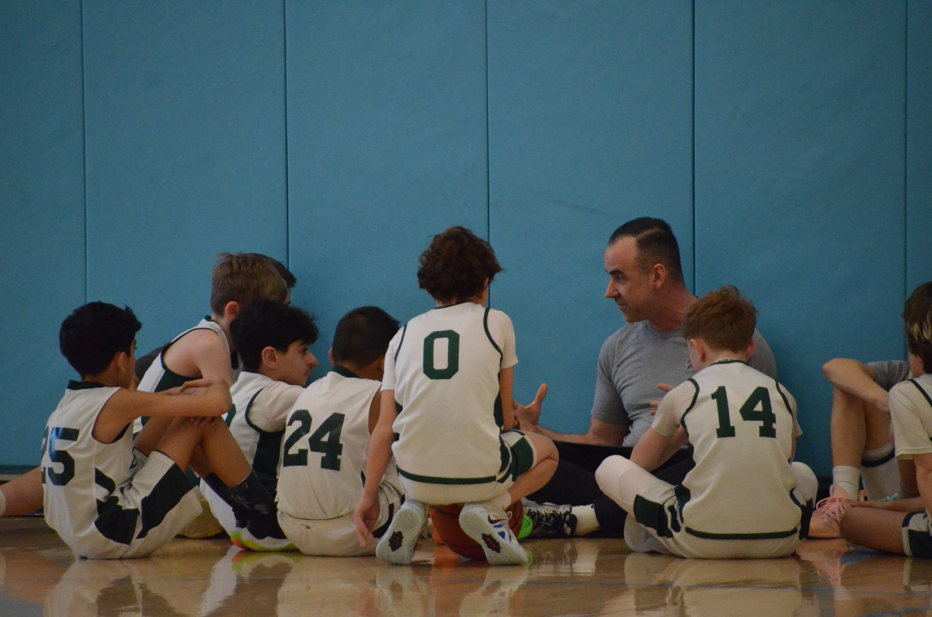 Youth basketball team sits on gym floor listening to a coach against a blue wall.