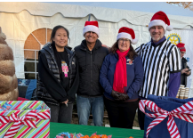 Four people in Santa hats stand behind wrapped gifts at a holiday table outdoors.