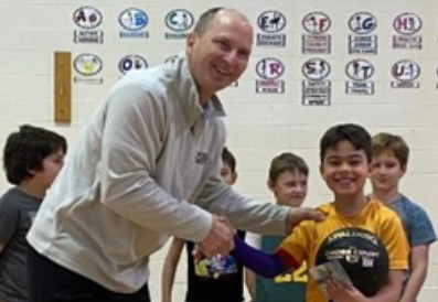 Man handing a medal to a smiling boy in a classroom with other children watching