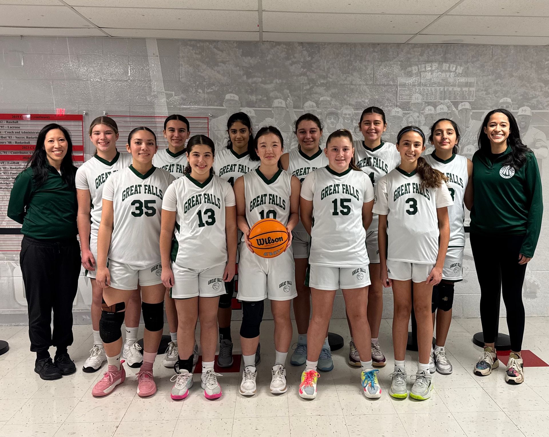 Youth basketball team in white uniforms posing indoors with coaches and a basketball center.