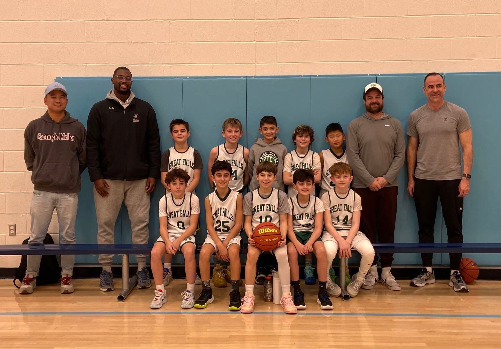 Youth basketball team posing in gym, with coaches standing behind them and a basketball held at center.