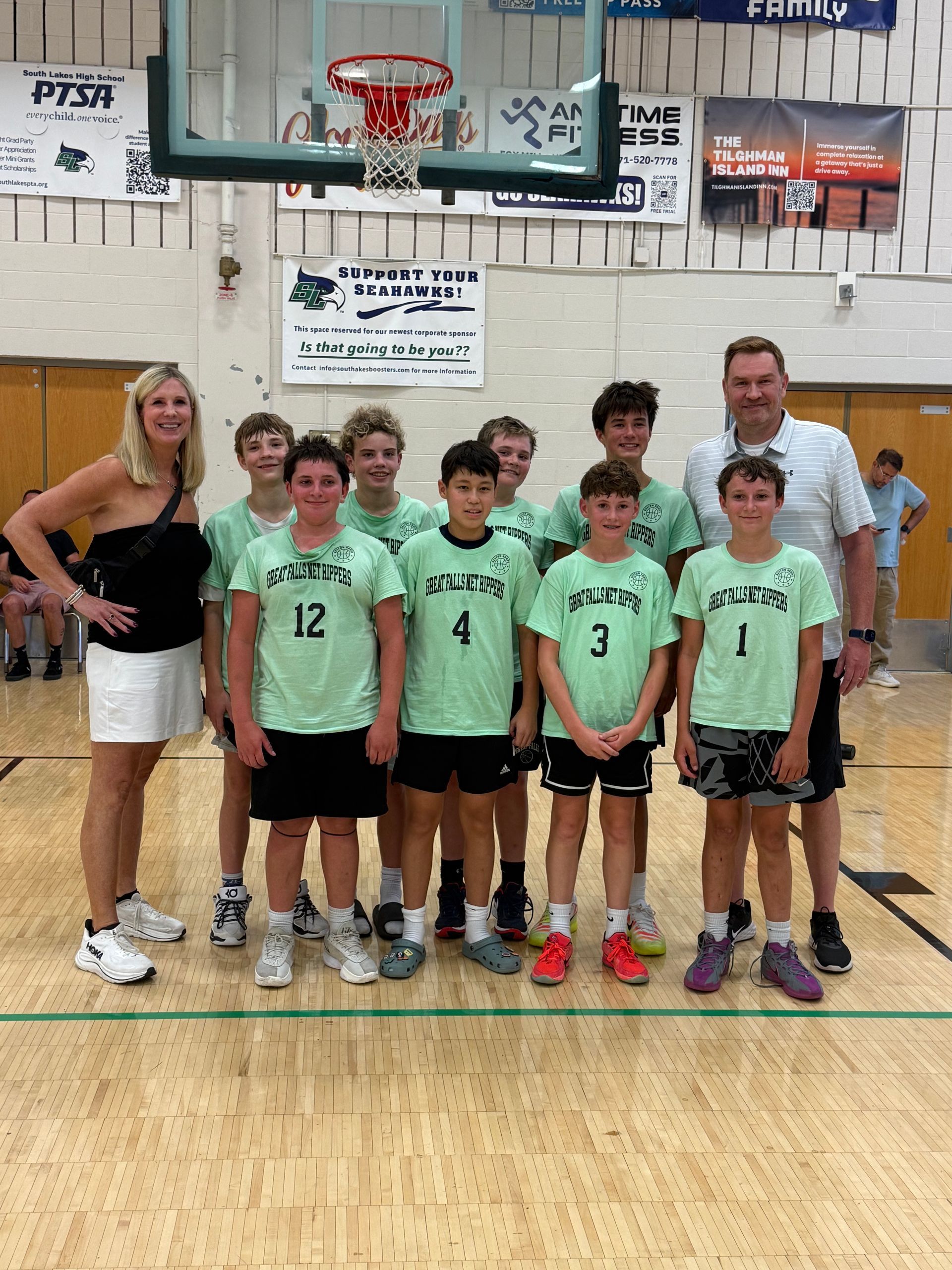 Youth basketball team posing with coaches in a gym, wearing mint green jerseys in front of the hoop.