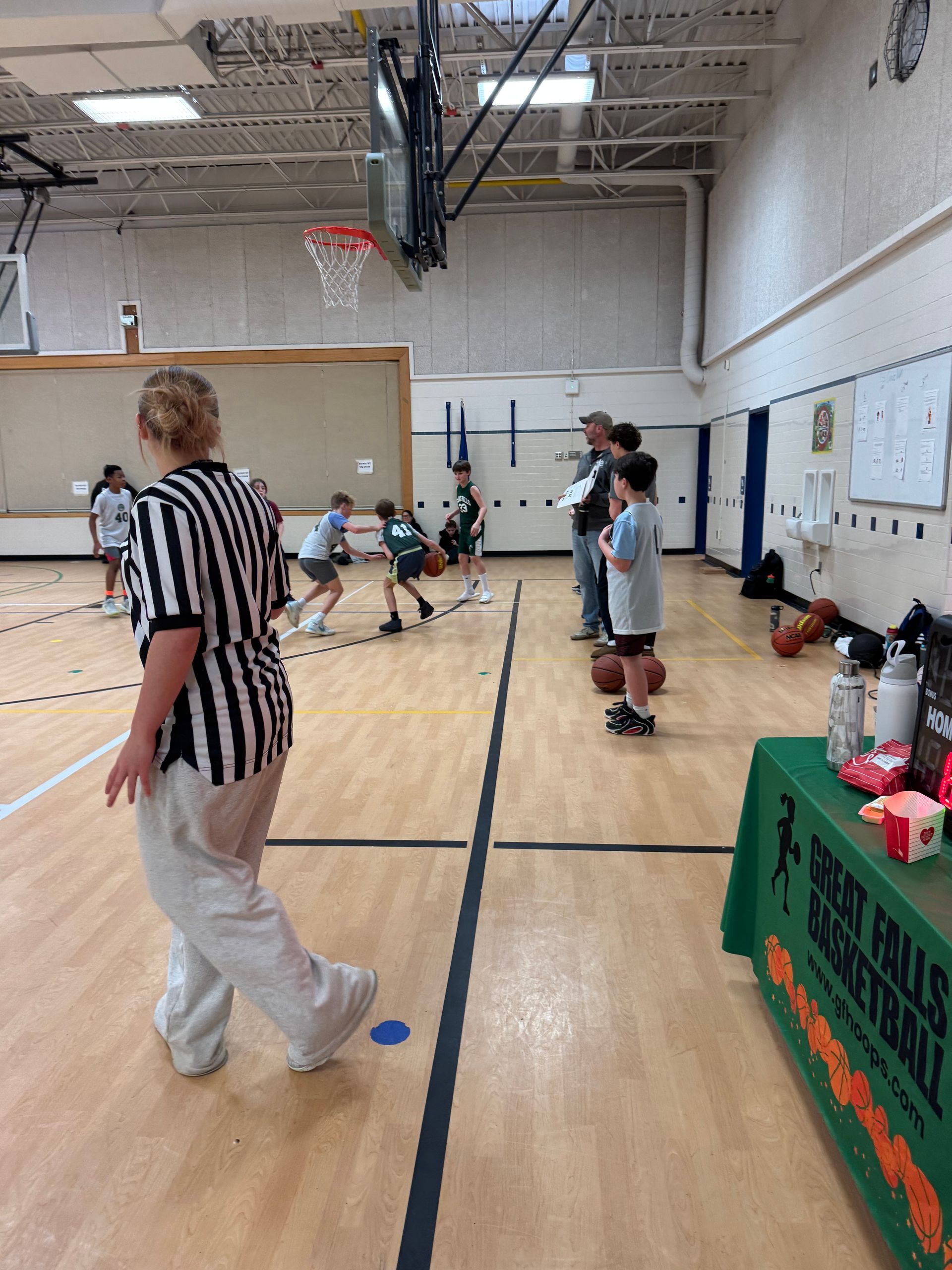 Indoor gym with a basketball hoop, children playing, and a referee near the court sideline