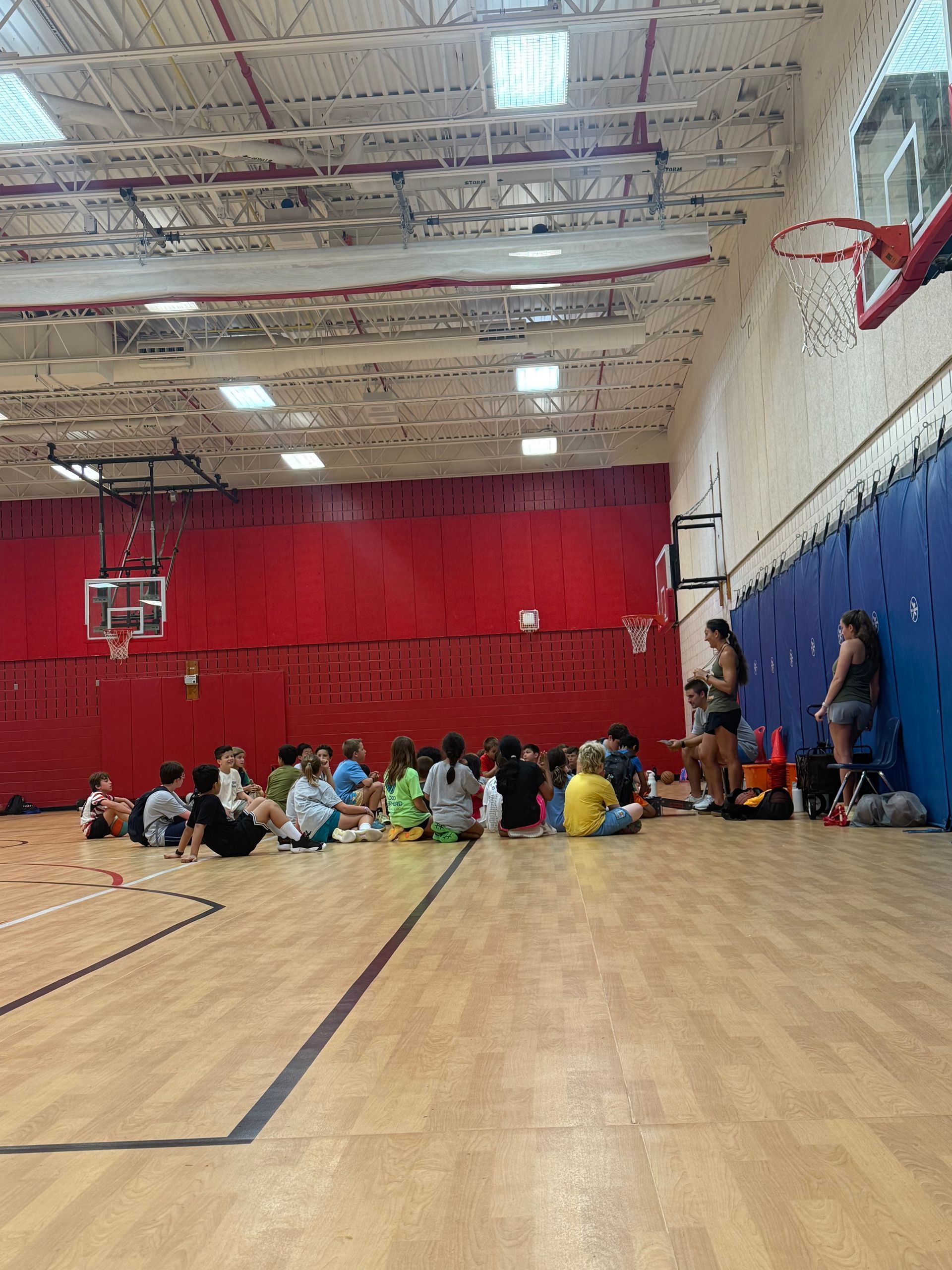 Children sitting in a gymnasium on a wooden court, with red and blue walls and basketball hoops overhead.