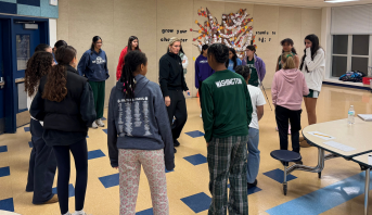 People standing in a circle in a classroom, facing an instructor near a wall mural.