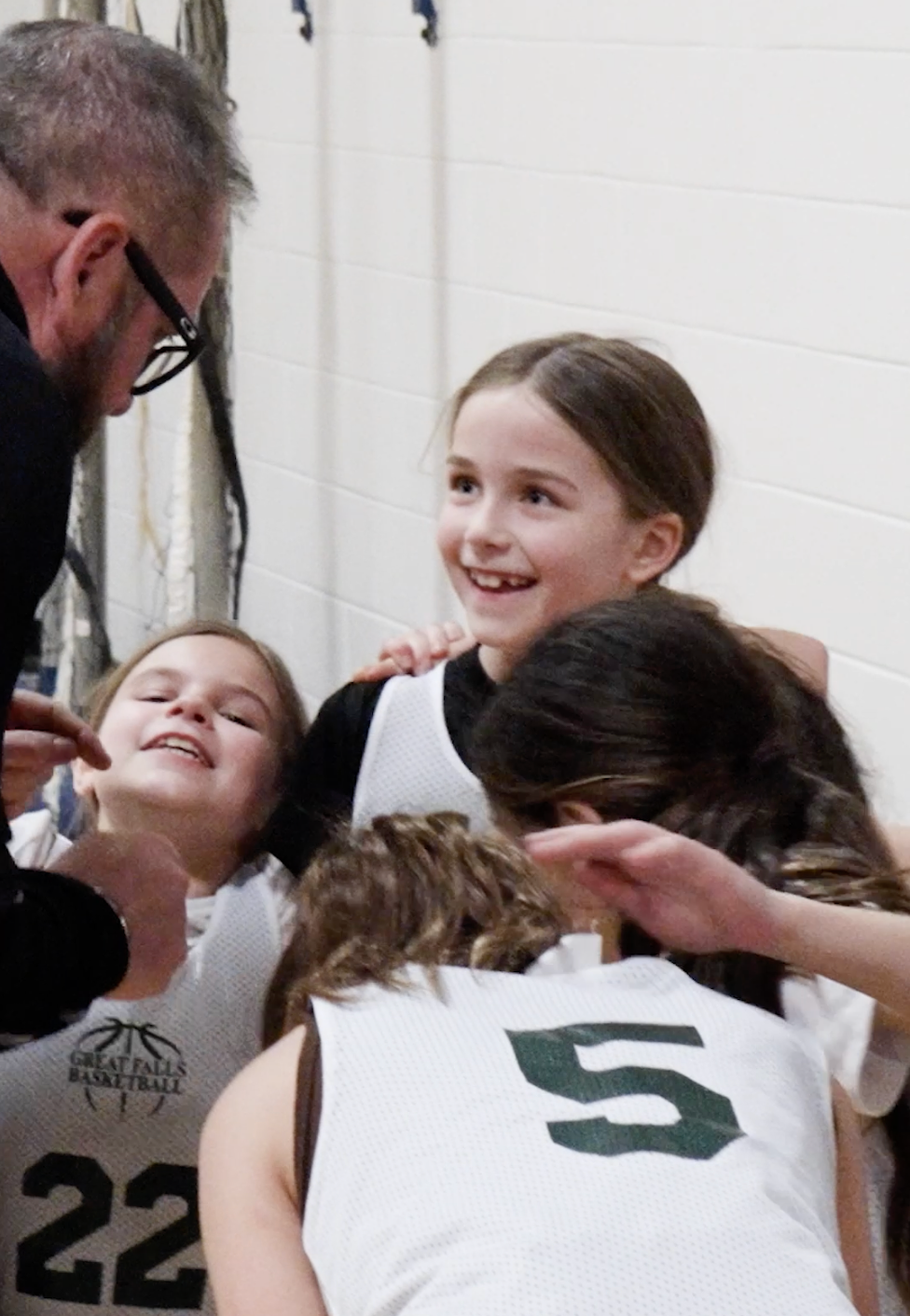 Girls in white sports jerseys huddle with a coach, smiling in a gym.
