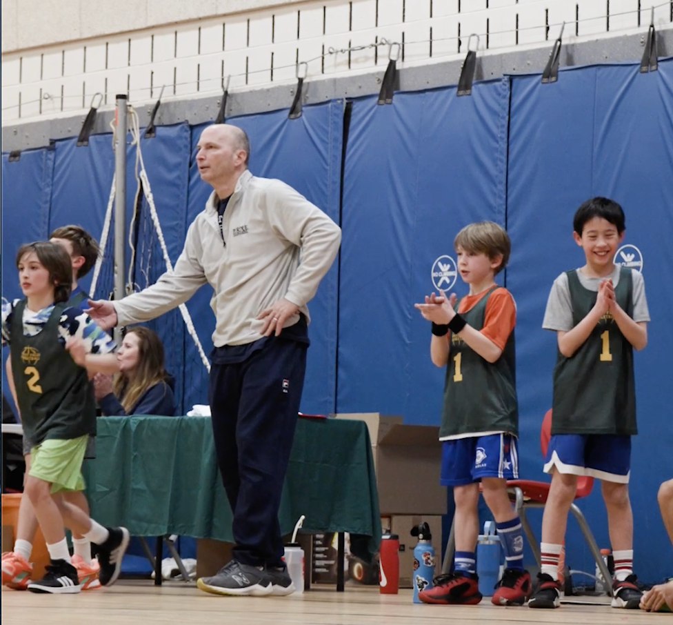 Coach stands beside young volleyball players on a gym court, with children in jerseys clapping near the bench.