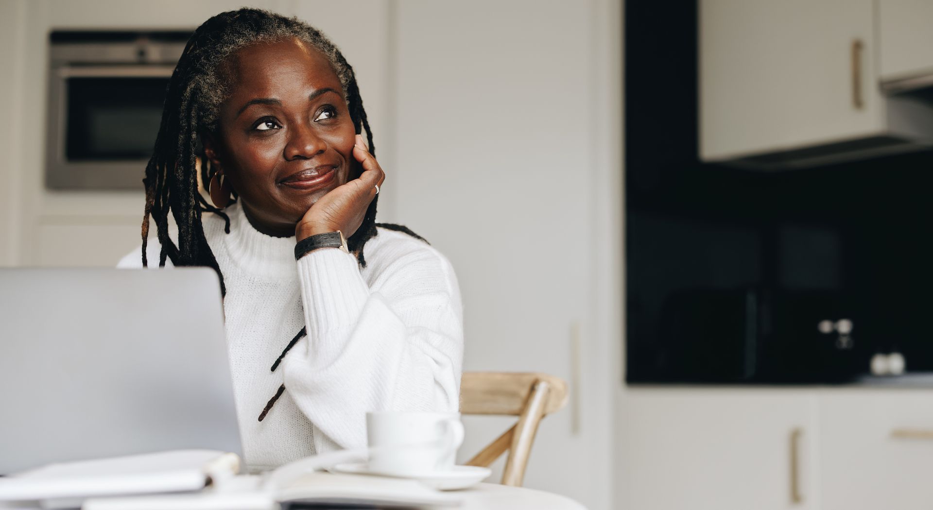 Woman with dreadlocks, resting chin on hand, looking up thoughtfully near laptop and coffee cup.