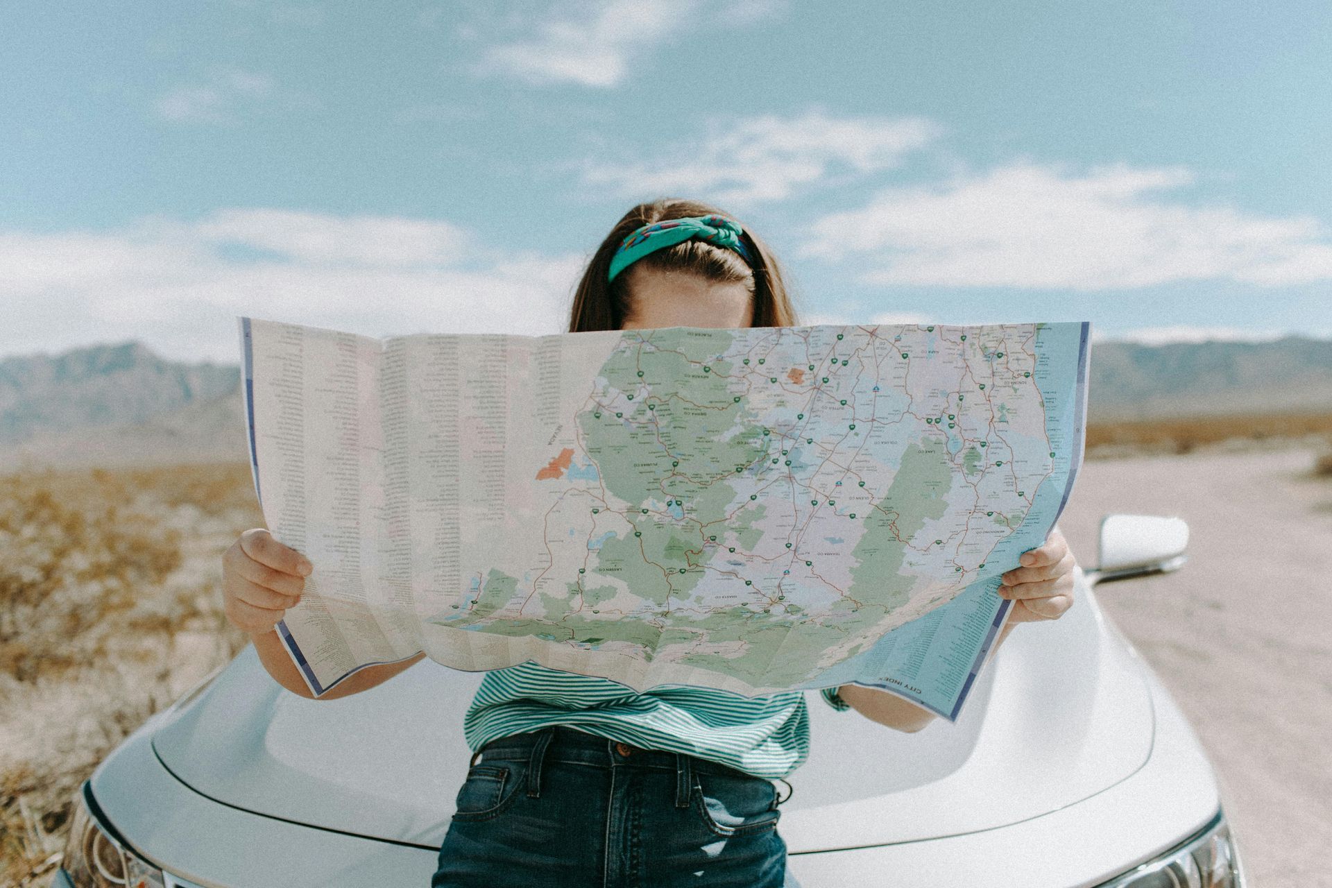 Person with a teal bandana over their eyes, holding a map, leaning on a car in a desert.