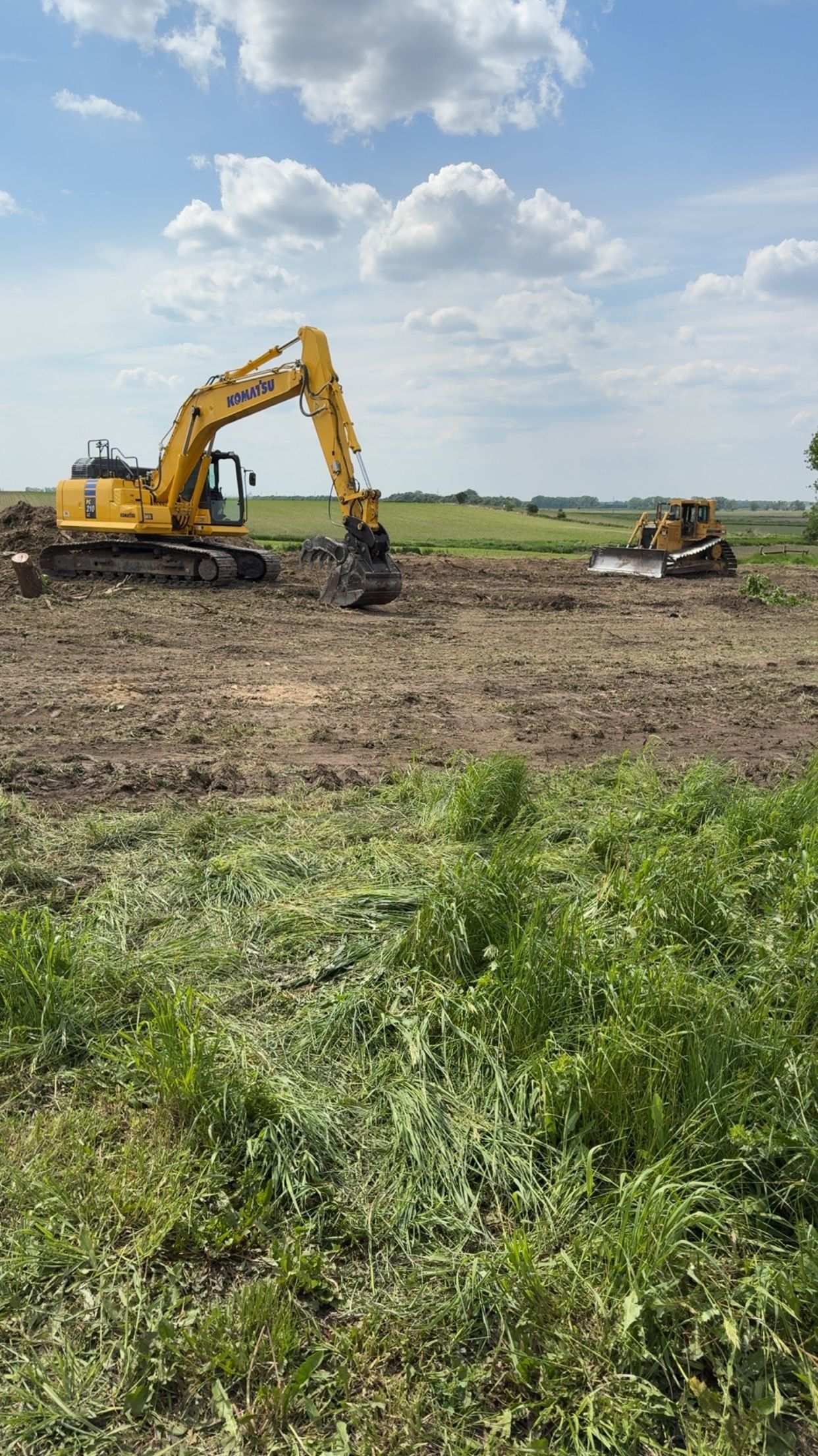 Yellow excavator and bulldozer working on a muddy field under a blue sky with clouds.