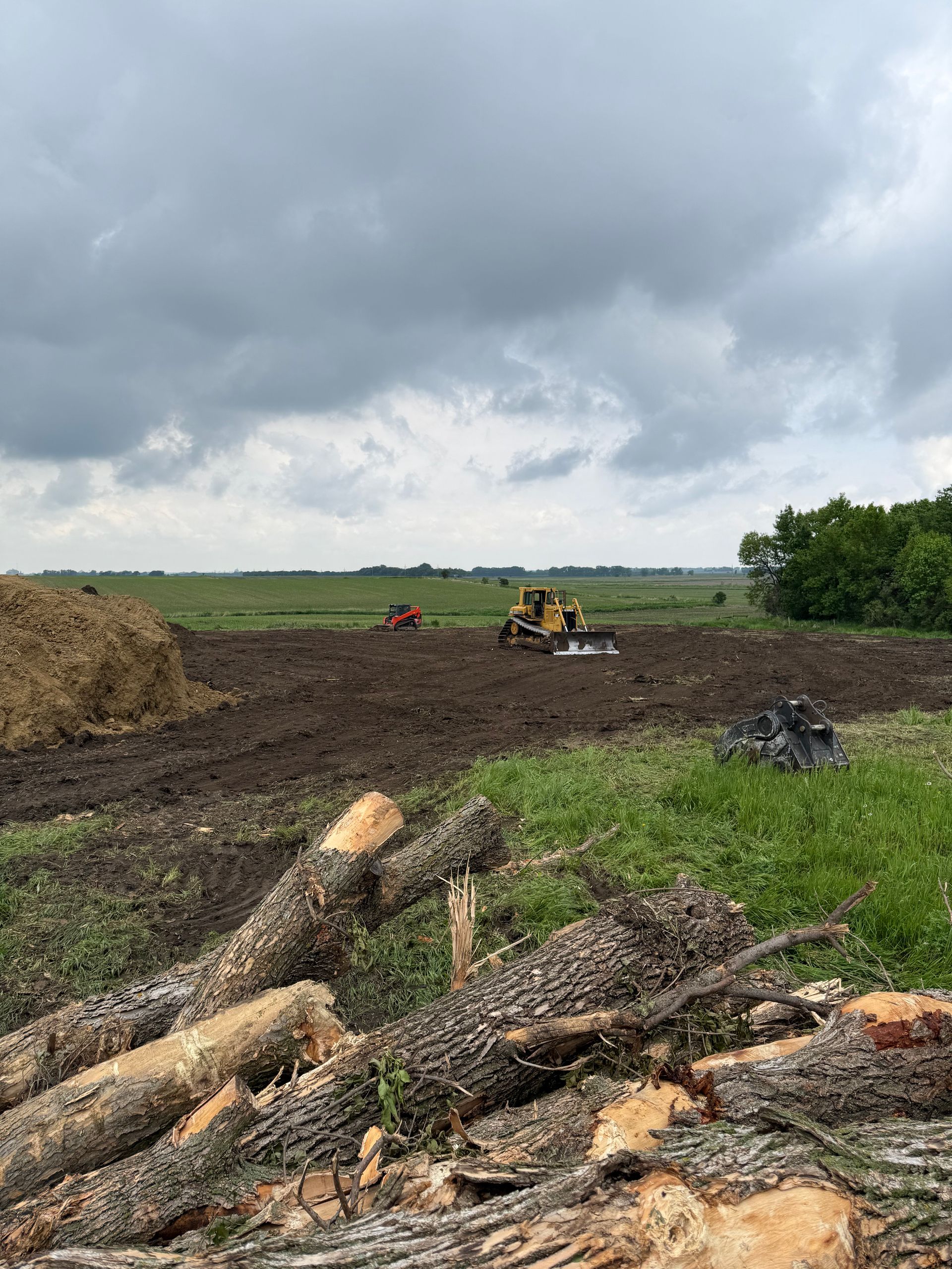 Bulldozers clearing a field under a cloudy sky, with logs in the foreground.